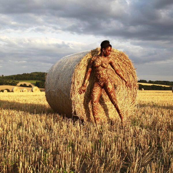 Für das Kunstprojekt AGRAR ART hat Bodypaint-Künstler Jörg Düsterwald mit Körperfarbe ein dunkelhäutiges Fotomodell mit goldfarbenen Ornamenten bemalt. Die Frau posiert in der Abendsonne bei Strohballen auf einem Strohfeld.