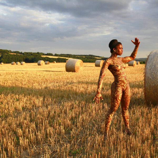 Für das Kunstprojekt AGRAR ART hat Bodypaint-Künstler Jörg Düsterwald mit Körperfarbe ein dunkelhäutiges Fotomodell mit goldfarbenen Ornamenten bemalt. Die Frau posiert in der Abendsonne bei Strohballen auf einem Strohfeld.