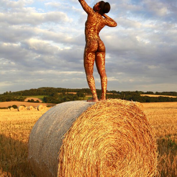 Für das Kunstprojekt AGRAR ART hat Bodypaint-Künstler Jörg Düsterwald mit Körperfarbe ein dunkelhäutiges Fotomodell mit goldfarbenen Ornamenten bemalt. Die Frau posiert in der Abendsonne bei Strohballen auf einem Strohfeld.