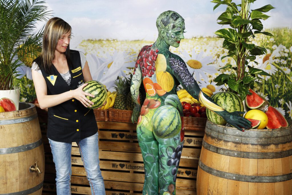 Für das Kunstprojekt WORKING ART, bei dem Berufe mit Bodypaintingmodellen dargestellt werden, hat Künstler Jörg Düsterwald ein Fotomodell vollständig bemalt. Die mit Obst und Früchten gestylte Frau hält sich zusammen mit einer Verkäuferin an einem Obststand auf.