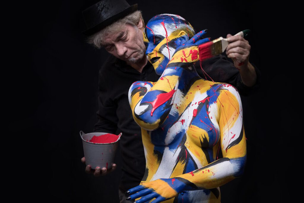 Eine nackte, von Künstler Jörg Düsterwald mit bunter Farbe versehene Frau steht in einem Fotostudio vor einer farbigen Kulisse. Der Künstler hält einen Pinsel in der Hand und besprenkelt sie mit Farbe.