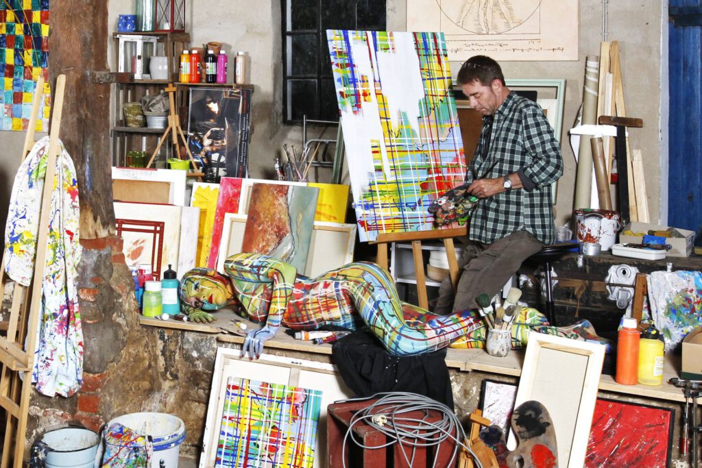 Künstler Jörg Düsterwald arbeitet in seinem Atelier an einem Gemälde-Bildwerk. Eine farbig bemaltes, nacktes Fotomodell leistet ihm Gesellschaft.