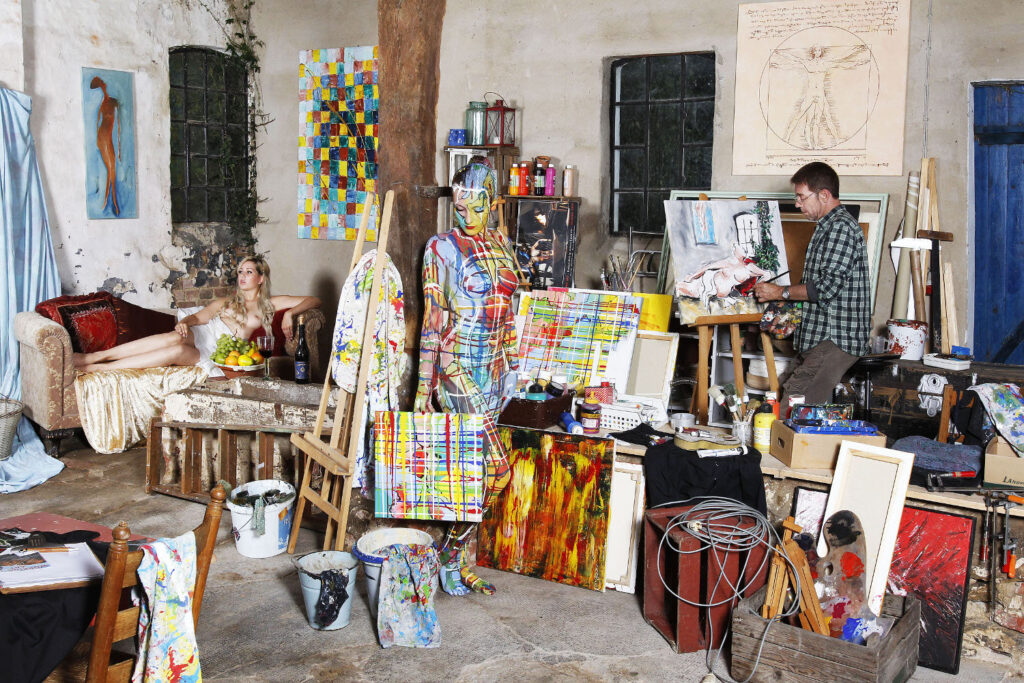 Künstler Jörg Düsterwald arbeitet in seinem Atelier an einem Gemälde-Bildwerk, bei dem eine Muse als Vorlage auf einem Sofa liegt. Eine farbig bemaltes, nacktes Fotomodell leistet den beiden Gesellschaft.