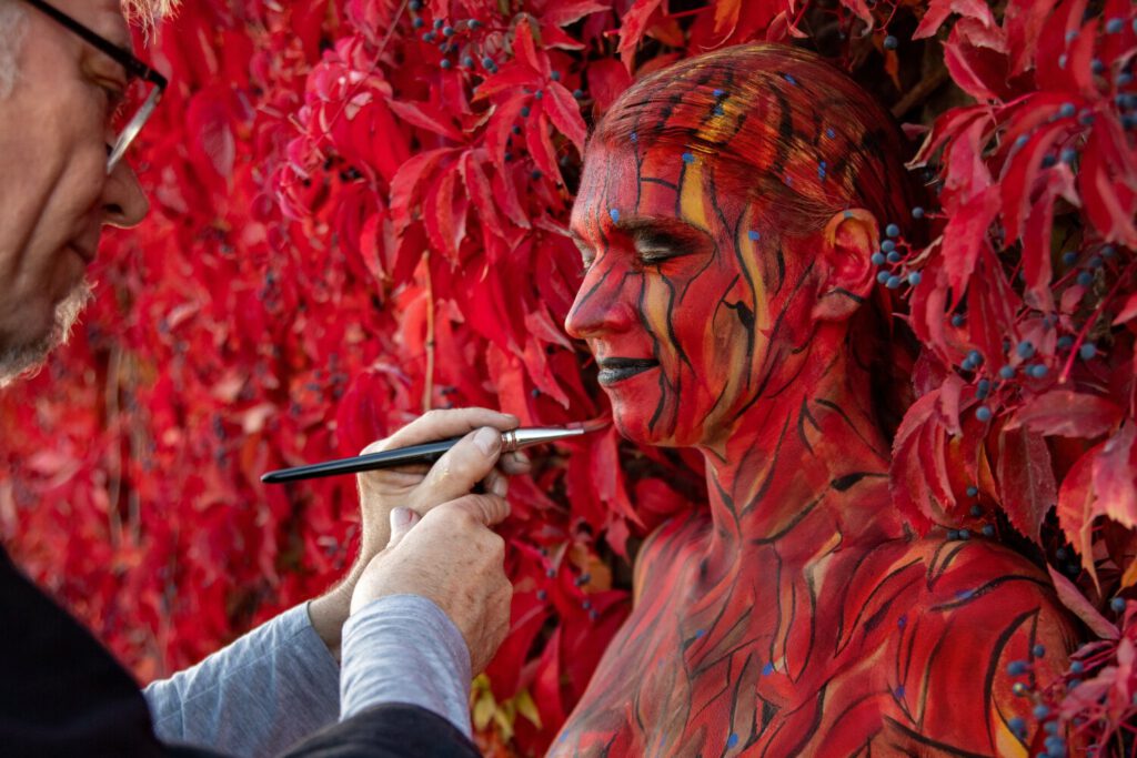 Ein nacktes Fotomodell steht zwischen den roten Blättern einer Weinrankenwand. Künstler Jörg Düsterwald hält einen Pinsel mit roter Farbe in der Hand und bemalt die Frau vollständig so mit Farbe, dass sie mit der Kulisse verschmilzt.